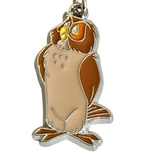 May include: A metal owl keychain with a silver border. The owl has a light brown body, dark brown wings, and a yellow beak. The owl is in a standing position, with its head turned slightly to the side.
