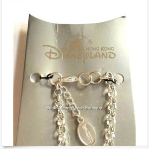 Puede incluir: Un brazalete de cadena de plata con un dije de plata que presenta el logotipo de Disneyland Hong Kong. El dije est&aacute; unido a la cadena con un cierre de plata. El brazalete se muestra sobre un fondo plateado con las palabras "Hong Kong Disneyland" impresas en &eacute;l.