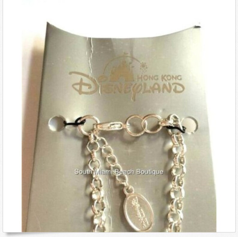 Puede incluir: Un brazalete de cadena de plata con un dije de plata que presenta el logotipo de Disneyland Hong Kong. El dije est&aacute; unido a la cadena con un cierre de plata. El brazalete se muestra sobre un fondo plateado con las palabras "Hong Kong Disneyland" impresas en &eacute;l.