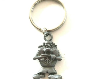 May include: A silver-colored metal keychain featuring a cartoonish figure holding a peace sign. The figure has a stern expression and is wearing a hat and tunic. The keychain ring is silver and attached to the figure's head.
