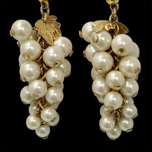 May include: A pair of vintage gold-tone earrings, each featuring a cluster of faux pearls resembling grapes. The earrings have a gold leaf accent at the top and are set against a black background.