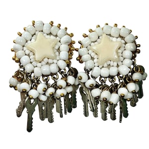 May include: A pair of statement earrings featuring a star design in the center, surrounded by white beads and gold accents. Dangling from the earrings are small silver keys and more white beads. The earrings have a unique, eye-catching design.