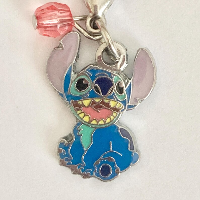 Puede incluir: Un charm azul y plateado con el personaje Stitch, con orejas rosas y una amplia sonrisa. Una cuenta rosa cuelga sobre el charm, unida a un cierre plateado. El charm es probablemente para una pulsera o collar.