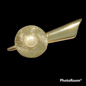 May include: A gold-tone metal brooch featuring a circular center with a radiating sunburst pattern and a smooth, off-white faux pearl cabochon at its core. A sleek, elongated, and tapered gold-tone metal wing extends from the right side of the circle.