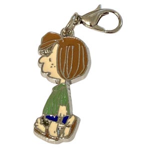 May include: A silver-toned charm featuring a cartoon character in profile. The character has brown hair and a brown hat, a green shirt, blue shorts, and cream-colored shoes. The charm is attached to a silver clasp.