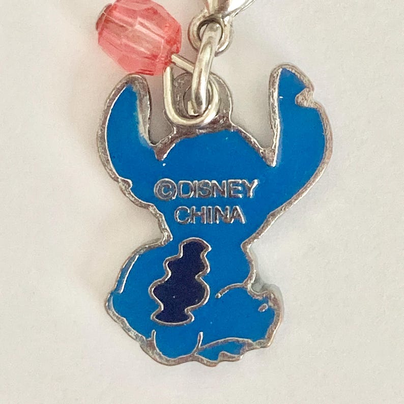 Puede incluir: Un dije de Stitch azul con un lazo de metal plateado y una cuenta rosa. El dije presenta el logotipo de Disney y el texto "CHINA". Un art&iacute;culo de colecci&oacute;n.