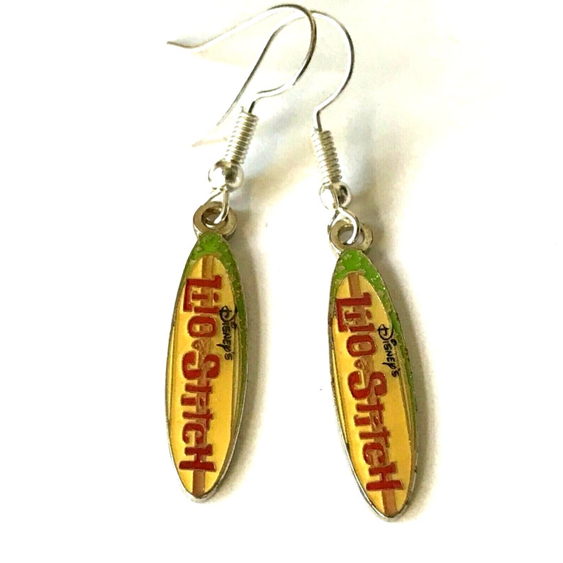May include: A pair of silver dangle earrings featuring a surfboard design with the text "Lilo & Stitch" and the Disney logo. The surfboard is yellow with red and green accents.