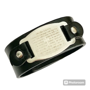 May include: A black leather bracelet with a silver metal plate engraved with a prayer. The prayer is in English and includes the words "God is love" and "forever Amen".