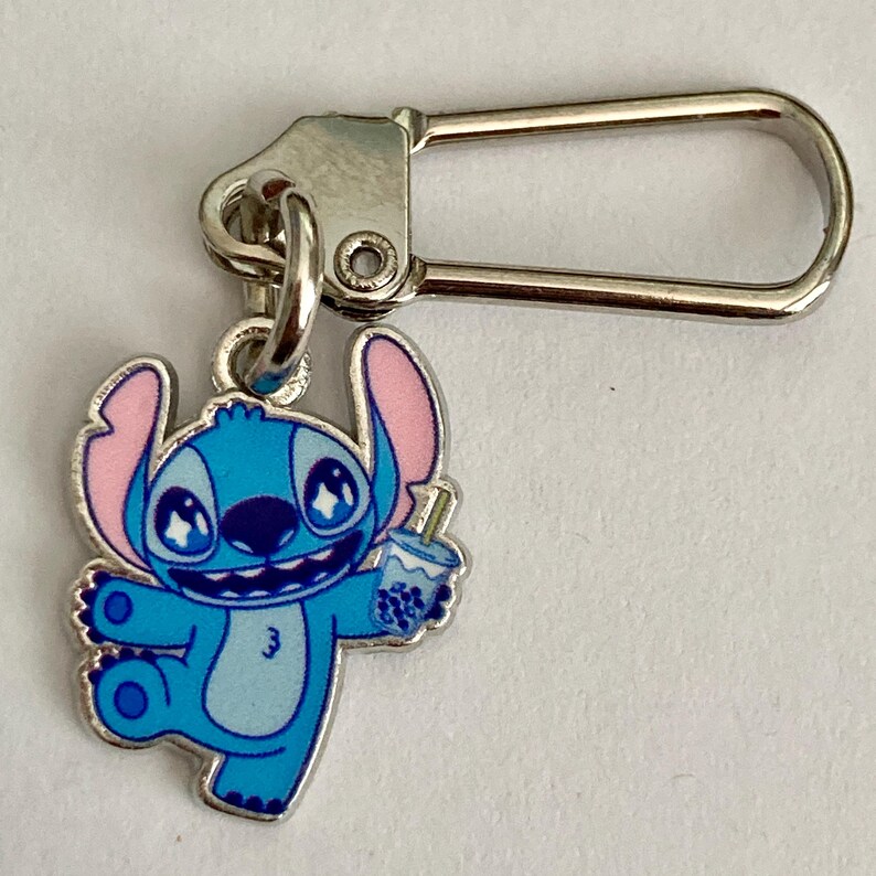 Puede incluir: Un charm de esmalte azul y rosa con el personaje Stitch, sosteniendo una bebida. El charm est&aacute; unido a un cierre de metal plateado, adecuado para un llavero o un accesorio de bolso.