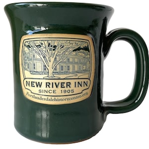 May include: A dark green ceramic mug with a curved handle. The mug features a rectangular label with an illustration of a building and a tree, with the text "NEW RIVER INN SINCE 1905" and a website address.