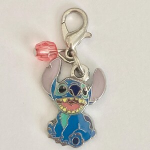Puede incluir: Un charm plateado con el personaje Stitch de Lilo & Stitch. El charm es azul con orejas rosas y una cara sonriente. Una cuenta rosa est&aacute; unida al charm con un cierre plateado.
