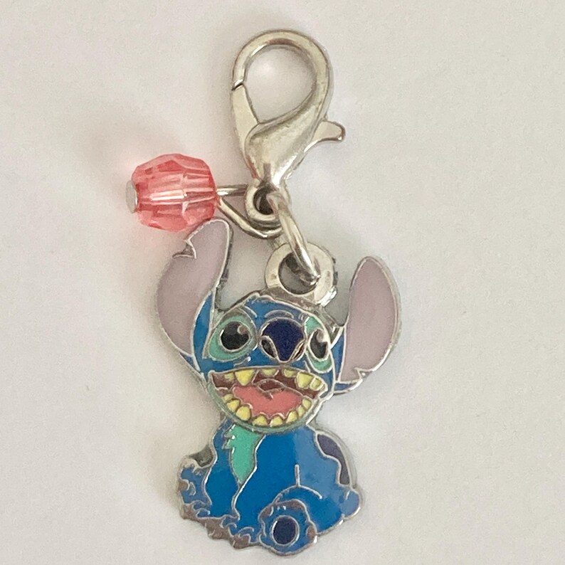 Puede incluir: Un charm plateado con el personaje Stitch de Lilo & Stitch. El charm es azul con orejas rosas y una cara sonriente. Una cuenta rosa est&aacute; unida al charm con un cierre plateado.