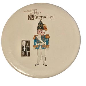 May include: A round, off-white button featuring a detailed illustration of a Nutcracker soldier in a blue uniform with gold accents. The text "The Nutcracker" and "Miami City Ballet" are also displayed.