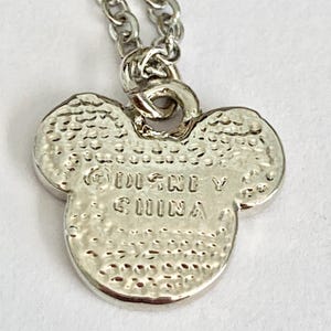K&ouml;nnte beinhalten: Silberne Halskette mit einem Anh&auml;nger in Form eines Mickey Mouse Kopfes. Auf der R&uuml;ckseite des Anh&auml;ngers ist der Text "Disney China" eingraviert.