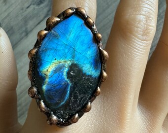 Puede incluir: Un anillo con una gran piedra preciosa azul de forma ovalada engastada en un engaste de metal de color cobre. La piedra preciosa presenta un patrón azul oscuro y negro con toques de oro.