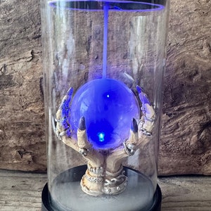 May include: A glass incense burner with a skeletal hand holding a glowing blue orb. The burner has a black base and a clear glass dome.