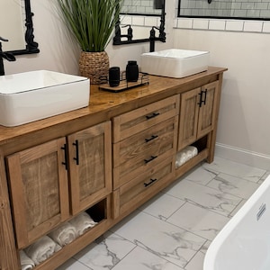 May include: A wooden bathroom vanity with two sinks, three drawers, and a shelf for towels. The vanity has a light brown wood finish and black hardware. The vanity is in front of a white tiled wall with a white marble floor.