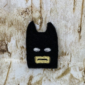 May include: A black felt Batman mask patch with white embroidered eyes and a gold embroidered mouth. The patch has two pointed ears and is set against a textured, light brown background.