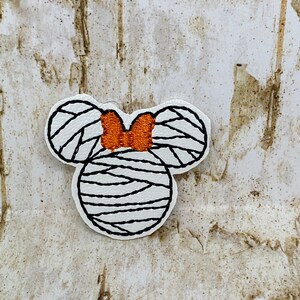 May include: A white patch in the shape of a mouse head with black lines and an orange bow. The mouse head is wrapped in black lines, resembling a mummy. The patch is on a light brown, textured background.