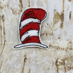 May include: A fabric patch featuring the iconic red and white striped hat from a children's book. The patch has a white border and is set against a textured, light-colored background.