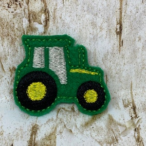 Könnte beinhalten: Ein grüner Filz-Traktor-Applikation mit schwarzen Rädern und gelben Zentren. Der Traktor hat eine silberne und grüne gestickte Kabine vor einem strukturierten, hellfarbigen Hintergrund. Dieser Aufnäher eignet sich zum Aufnähen oder Aufbügeln auf Stoff.