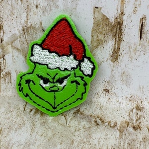 Op de afbeelding: Een groen geborduurd embleem met het gezicht van de Grinch, met een rode kerstmuts met witte rand. De patch heeft een zwarte omtrek en is geplaatst tegen een getextureerde, gebroken witte achtergrond.