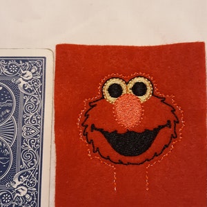 May include: A red felt square featuring an embroidered Elmo face with gold, black, and orange details. A blue playing card is next to the felt square.