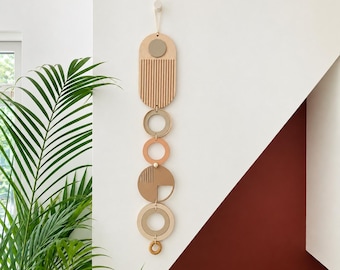 May include: A vertical wall hanging composed of interconnected wooden shapes in shades of beige, tan, and terracotta. The piece features an elongated arch with vertical grooves, several circular rings, and a solid semi-circle with line details.