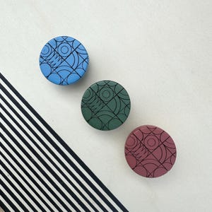 Può includere: Tre oggetti decorativi rotondi e colorati. Uno è blu, uno verde e uno rosso tenue. Ognuno ha un motivo geometrico nero. Gli oggetti sono disposti su una superficie bianca con un accento diagonale a strisce bianche e nere.