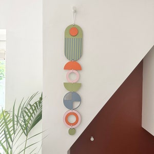 May include: A wall hanging made of wood with geometric shapes in shades of green, blue, pink, and orange. The shapes are arranged vertically and connected by string.