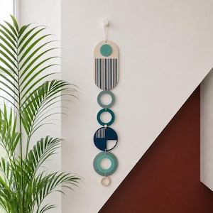 Pode incluir: Um pendente de parede vertical composto por formas geométricas interligadas em tons de azul-petróleo, azul-marinho e madeira clara. A peça apresenta um retângulo arredondado superior com listras verticais, seguido por vários anéis e um círculo.
