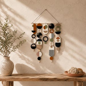May include: A wall hanging featuring five vertical strands of geometric shapes suspended from a horizontal wooden rod. The shapes include circles, semi-circles, and rounded rectangles in a palette of black, white, olive green, and terracotta orange.