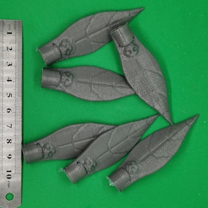 Peut inclure: Six objets gris en forme de feuilles, avec une surface texturée et une base cylindrique, sont disposés sur un fond vert. Une règle métrique est à côté. Chaque feuille mesure environ 10 cm de long.