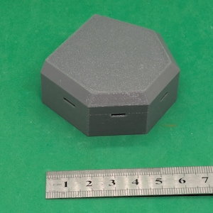 Può includere: Una scatola grigia di forma geometrica con superficie strutturata e piccole aperture rettangolari. Un righello di metallo con misure in centimetri è posizionato sotto la scatola.