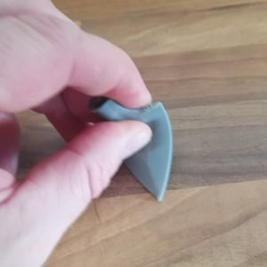 Peut inclure: Un médiator de guitare gris avec un dessus arrondi et un bas pointu. Le médiator est tenu entre les doigts contre une surface en bois. Le médiator a un bord texturé.