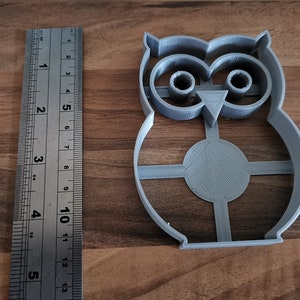 Pu&ograve; includere: Un tagliabiscotti a forma di gufo in plastica grigia con un righello accanto. Il tagliabiscotti &egrave; alto circa 10 cm. Il righello mostra le misure in pollici. Il tagliabiscotti ha un corpo circolare con un design a croce.