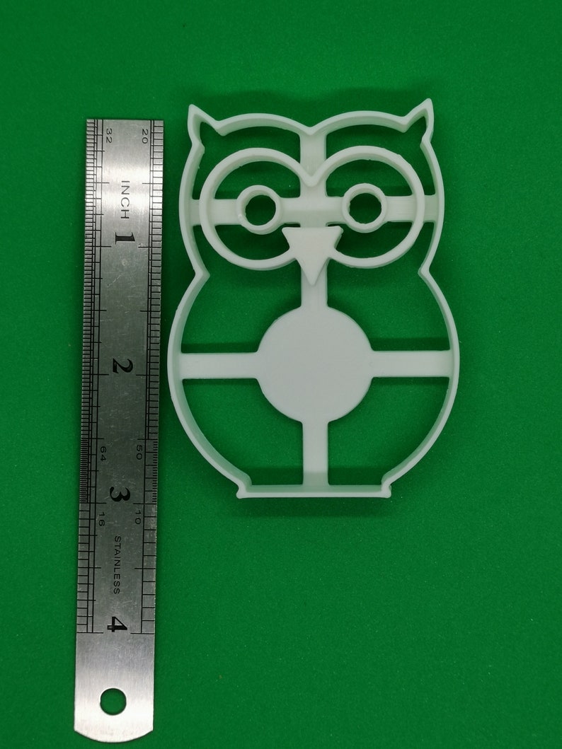 Pu&ograve; includere: Un tagliabiscotti a forma di gufo in plastica bianca con un righello accanto. Il tagliabiscotti &egrave; alto circa 10 cm. Il design del gufo presenta grandi occhi, un becco e un corpo rotondo. Lo sfondo &egrave; di colore verde.