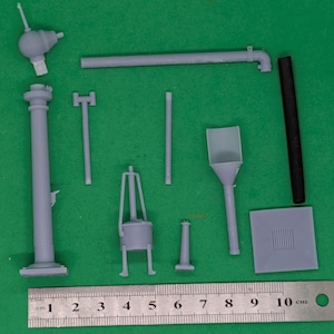 May include: A selection of grey plastic model components, including pipes, a tall column, and a small furnace, arranged on a green surface. A metal ruler with centimetre markings is at the bottom. The parts appear to be for a model kit.