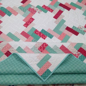 May include: A quilt featuring a white background with a repeating herringbone pattern made of rectangular fabric pieces in shades of pink, red, and teal. The quilt has a teal backing with a white polka dot pattern and visible quilting stitches.