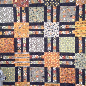 May include: A patchwork quilt featuring a grid of squares with Halloween-themed patterns including pumpkins, ghosts, and cats in orange, black, gray, and yellow. The fabric blocks are separated by black sashing and feature the word HALLOWEEN.