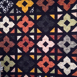 May include: A quilt featuring a repeating pattern of four-pointed star shapes in various colors including orange, yellow, black, and cream. The shapes are set against a solid black background with small triangular accents in the corners.