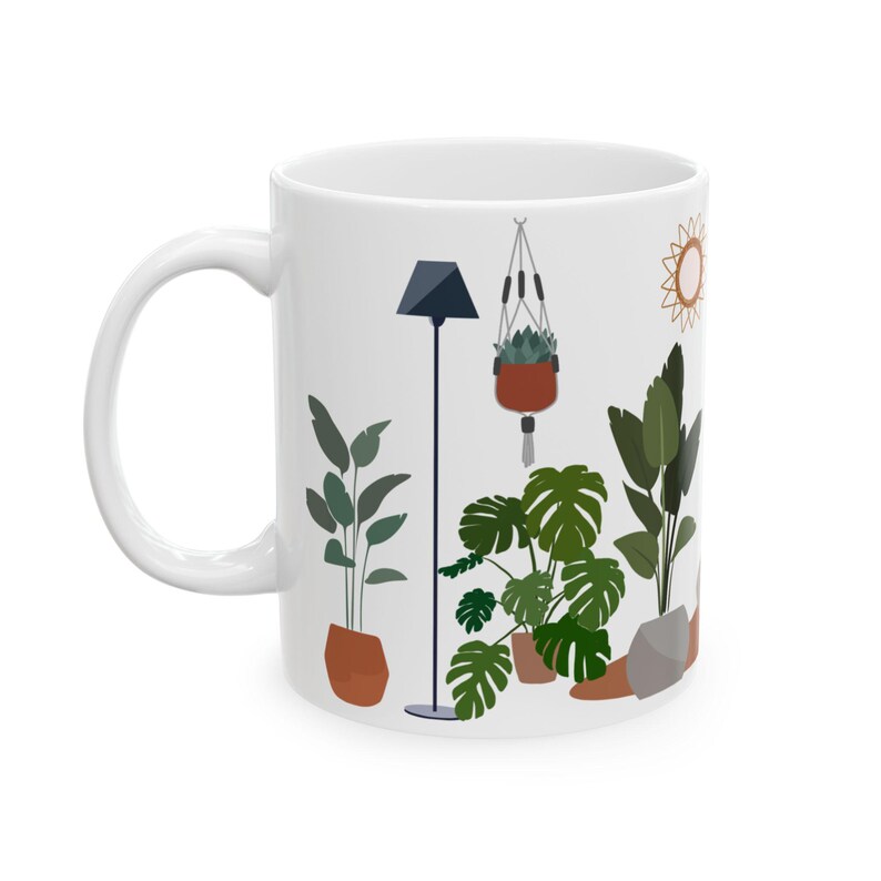 Pu&ograve; includere: Tazza in ceramica bianca con un'illustrazione colorata di piante da interno, una pianta sospesa, una lampada e un sole. Le piante sono in vasi di terracotta.