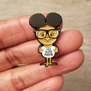 Könnte beinhalten: Ein schwarzer, goldener und weißer Emaille-Pin mit einem Cartoon-Küken mit schwarzen Afro-Puffs, das eine Brille und ein weißes T-Shirt mit den Worten "DOPE BLACK CHICK" trägt. Das Küken hat gelbe Füße und einen gelben Schnabel.