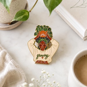 May include: A gold-toned enamel pin featuring a person with dark skin and curly hair wearing a green headband and a cream-colored sweater. The person holds a terracotta-colored pot with a green plant above their head. The word growing is written in green cursive script with a small heart below it.