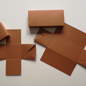 Könnte beinhalten: Vier braune Pappkartons, zwei sind flach gefaltet, einer ist teilweise gefaltet und einer ist vollständig zusammengebaut. Die Kartons sind alle gleich groß und haben ein schlichtes Design.