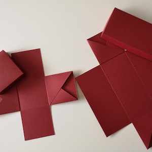 Könnte beinhalten: Burgunderfarbene Geschenkboxen in verschiedenen Stadien der Montage. Einige sind fertig montiert, andere teilweise gefaltet, was den Prozess zeigt. Die Boxen bestehen aus stabilem Papier oder Karton, ideal für kleine Geschenke.
