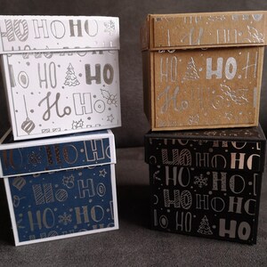 Könnte beinhalten: Vier kleine quadratische Geschenkboxen mit einem "Ho Ho Ho"-Muster. Die Boxen sind weiß, braun, blau und schwarz mit silbernen Folienbuchstaben.