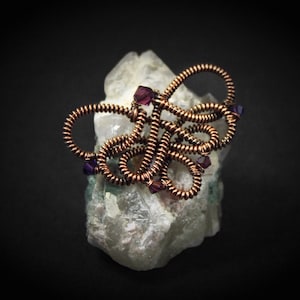 Op de afbeelding: Een sculptuur van koperdraad met paarse kralen, die lijkt op een bloem, rustend op een wit kristal.