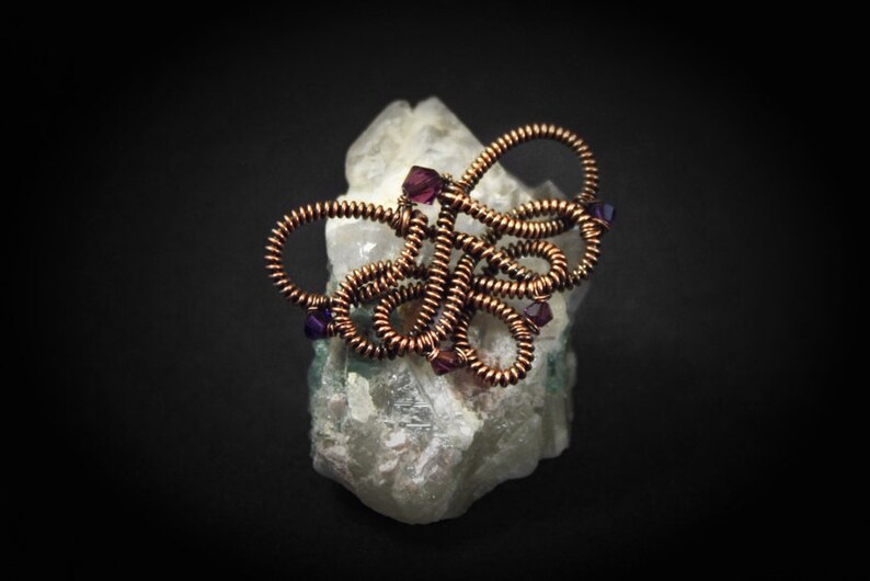 Op de afbeelding: Een sculptuur van koperdraad met paarse kralen, die lijkt op een bloem, rustend op een wit kristal.