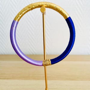 Può includere: Un braccialetto rigido in metallo dorato con sezioni di filo dorato, viola e blu. Il braccialetto è esposto su un supporto dorato su uno sfondo neutro. Il braccialetto è un accessorio di moda.